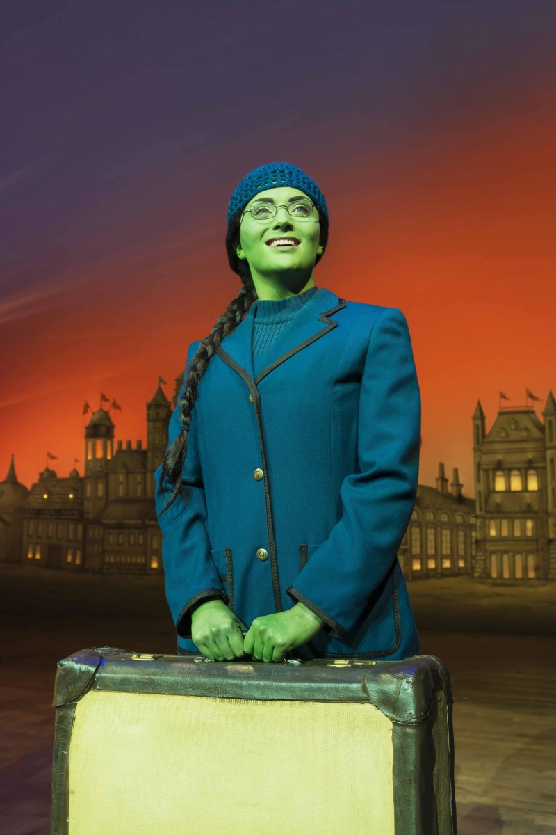 Elphaba smiling looking up holding suitcase in blue blazer